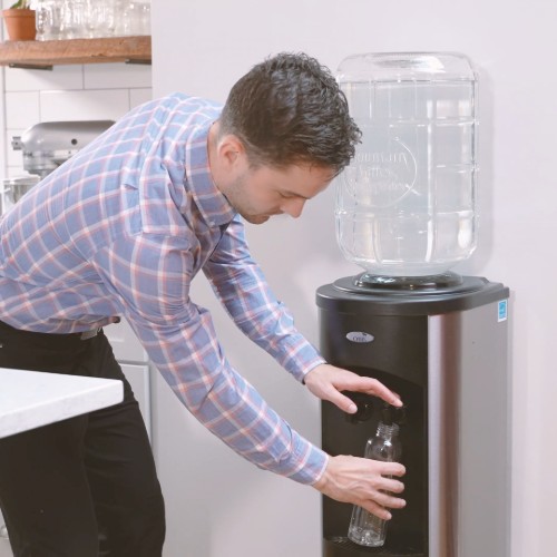 man in dress clothes filling a clear water bottle from a stainless steel water cooler with a mountain valley spring water glass bottle on top