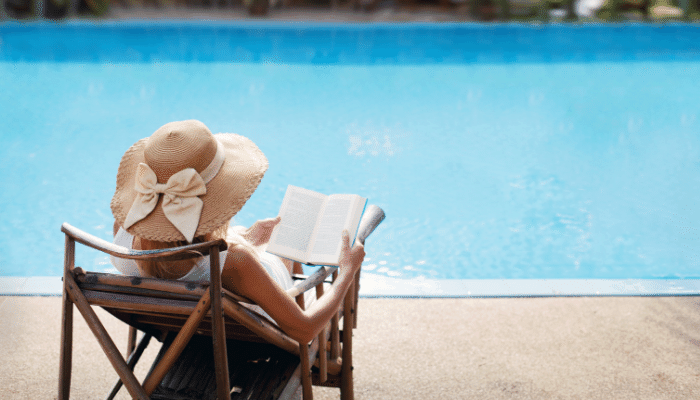 A person sitting in a lounge chair by a pool, reading a book and wearing a large straw hat.