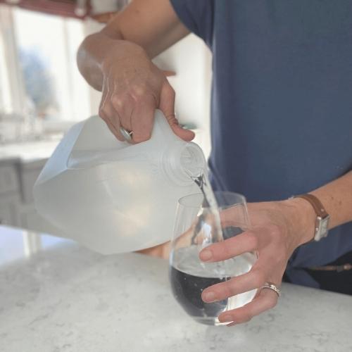 person in blue shirt pouring water from a one gallon jug into a clear glass on a white quartz counter top