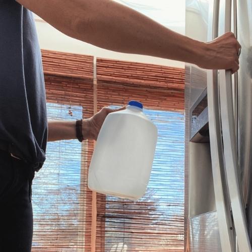 person holding a one gallon bottle of water with one hand and opening a refrigerator door with the other