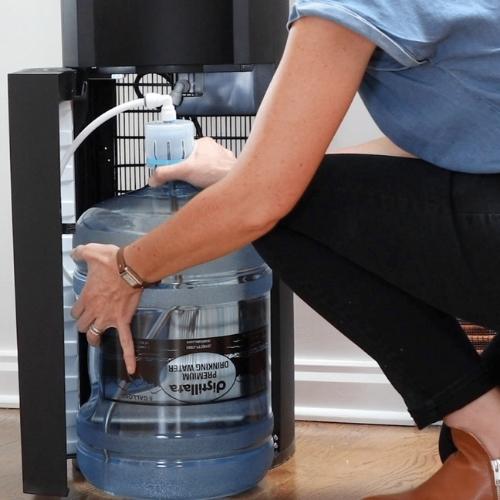 person bent down on one knee to place a distillata 5-gallon water bottle into a bottom load water cooler