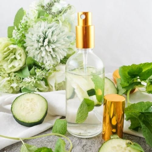 clear spray bottle with a gold nozzle sitting on a table surrounded by cucumber, mint, and flowers