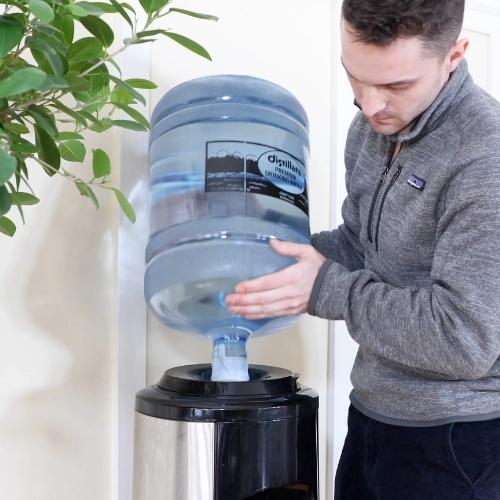 male in gray sweatshirt loading a 5-gallon bottle of distillata water onto a stainless steel water dispenser