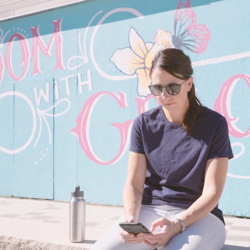 person sitting next to a reusabel water bottle on a ledge with a mural in the background while using a cell phone