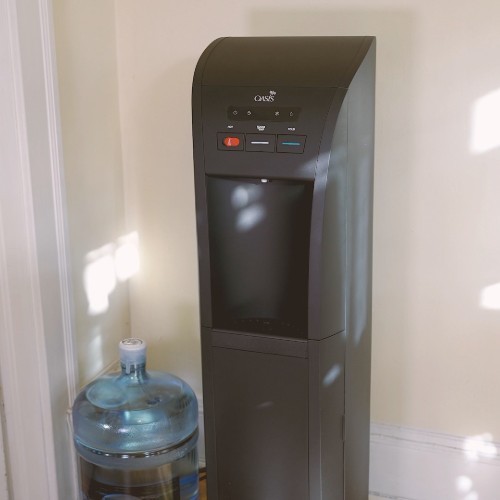 black bottom load water cooler sitting in a room with cream walls and white base board next to a 5-gallon bottle of water