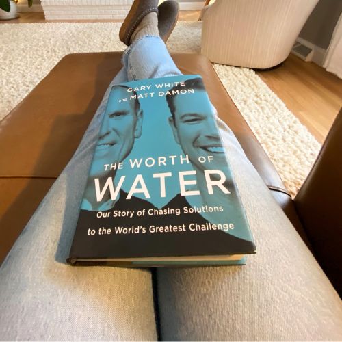 Person sitting with legs straight out on a couch with a copy of the book, "The Worth of Water" laying on lap.