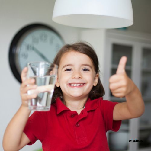 child in a red shirt holding a clear glass filled with water, a large clock is in the background