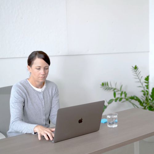 A woman with dark hair pulled back sits at a desk, focused on a gray laptop. A glass of water and a blue sticky note are to the right of the laptop on the light brown desk. A potted plant is visible in the blurred background against a white wall.